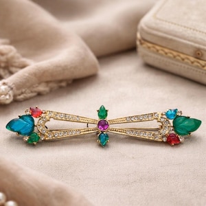 May include: A gold-toned brooch in a dragonfly design, adorned with colorful gemstones. The brooch features a central bar with clear crystals and is accented with blue, green, red, and purple stones. The wings are detailed with various jewel-toned gems.