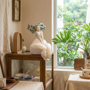 以下が含まれることがあります： 白いセラミック彫刻、座っている女性の像。なびく髪が特徴で、穏やかな表情をしています。木製のディスプレイキャビネットの上に置かれています。女性の髪は緑と青の混合色です。