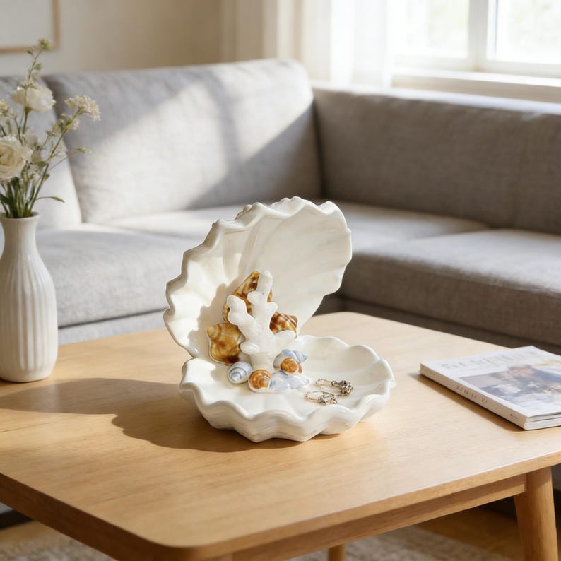 May include: A white ceramic seashell-shaped decorative dish. The dish is open, revealing a coral-like structure and several seashells. Small rings are placed in the dish. The dish is on a wooden table.