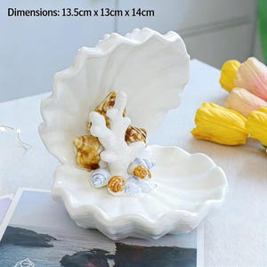 May include: A white ceramic seashell-shaped dish, 13.5 cm x 13 cm x 14 cm, with a ruffled edge. It features a coral and seashell design in the center. The dish is decorated with brown, white, and blue accents. The overall theme is coastal decor.