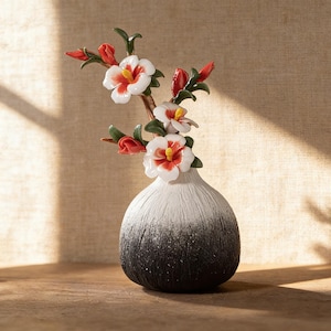 Könnte beinhalten: Eine Keramikvase mit einem Weiß-Schwarz-Verlauf hält ein Blumenarrangement. Das Arrangement zeigt weiße Blüten mit roten und gelben Zentren, rote Knospen und grüne Blätter. Die Vase hat eine strukturierte Oberfläche.
