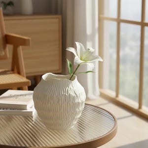 May include: A cream-colored, textured ceramic vase with a white lily flower. The vase has a unique, ridged design and sits on a round table. The flower has a green stem and leaves. The scene is set in a well-lit room.