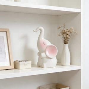 May include: A white ceramic elephant figurine with pink accents on the ears and cheeks. The elephant is sitting on a white shelf, next to a small white vase with dried flowers and a small beige box.