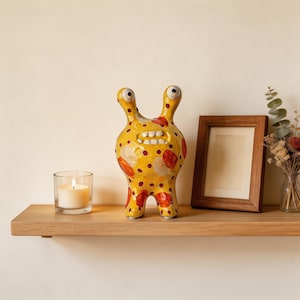 May include: A whimsical, yellow ceramic figurine with red and white accents and a toothy grin. The quirky creature has two large, upward-pointing eyes and is decorated with red polka dots. It sits on a wooden shelf with a candle and a framed picture.