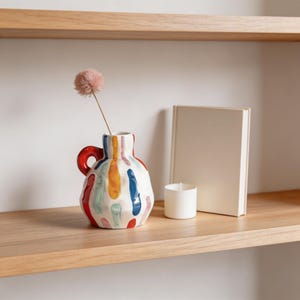 May include: A colorful ceramic vase with a red handle and vertical brushstrokes in various colors. A single pink dried flower is in the vase. A white candle and a blank book are on the wooden shelf.