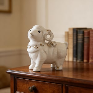 May include: A white ceramic ram ornament with gold detailing on the horns, hooves, and floral embellishments. The ram is positioned on a wooden surface. The figurine is a decorative item with intricate design.