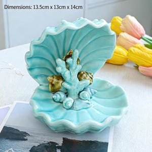 May include: A turquoise ceramic seashell-shaped dish with a coral and seashell centerpiece. The dish measures approximately 5.3 inches long, 5.1 inches wide, and 5.5 inches high. The dish is a decorative item.