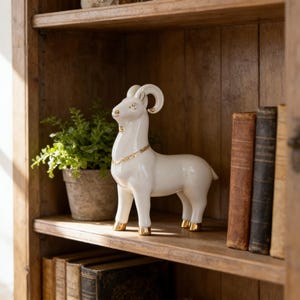 Könnte beinhalten: Eine weiße Keramik-Widderfigur mit goldenen Akzenten an Hörnern, Hufen und Halskette. Der Widder steht auf einem Holzregal neben einer Topfpflanze und mehreren antiken Büchern. Ein dekoratives Wohnaccessoire.