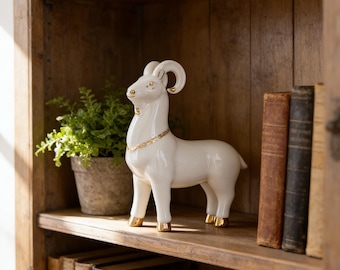 May include: A white ceramic ram figurine with gold accents on the horns, hooves, and a necklace. The ram is standing on a wooden shelf, next to a potted plant and several antique books. The figurine is a decorative home accent.