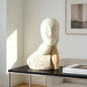 Könnte beinhalten: Eine cremefarbene Skulptur eines menschlichen Kopfes und Oberkörpers, die auf einer Hand ruht. Die Skulptur hat ein glattes, minimalistisches Design mit einer neutralen Farbpalette. Die Skulptur wird auf einem schwarzen Tisch ausgestellt.