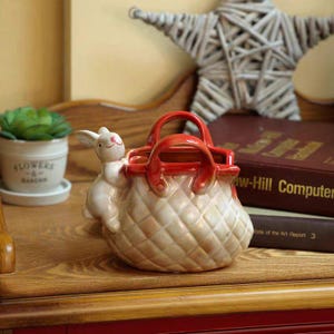 May include: A ceramic basket-shaped container with a quilted pattern, a red handle, and a white bunny figurine. The bunny is perched on the side of the basket. The container is tan and cream colored.