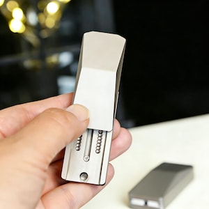 May include: A silver-colored metal toothpick holder with a sliding lid, held open to reveal the toothpicks inside. A second, closed holder sits on a white surface. The design is sleek and modern.
