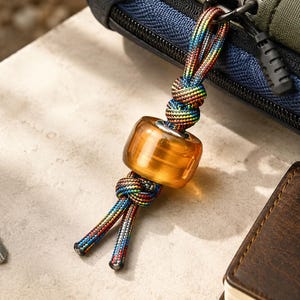 Op de afbeelding: Een sleutelhangeraccessoire met een doorschijnende, amberkleurige cilindrische kraal geregen aan een meerkleurig regenboog-paracord. Het koord is voorzien van decoratieve knopen boven en onder de kraal voor een stevig geheel.