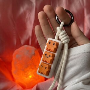Peut inclure: Un porte-clés composé d'une base rectangulaire blanche supportant trois touches de clavier carrées orange avec des visages pixelisés. Les touches sont alignées verticalement avec des expressions différentes. Un cordon tressé blanc relie le tout.