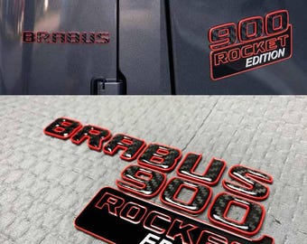 Può includere: Emblemi rossi e neri "BRABUS" e "900 ROCKET EDITION". Gli emblemi sono esposti su una superficie grigia del veicolo e su una superficie grigia strutturata. Il testo "900" ha un aspetto in fibra di carbonio.