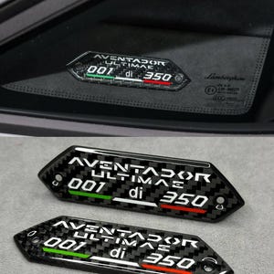 Może przedstawiać: Odznaka samochodowa z włókna węglowego z białym napisem "AVENTADOR ULTIMAE 001 di 350". Odznaka ma zielone i czerwone paski. Odznaka jest pokazana na szybie samochodu, a także osobno.