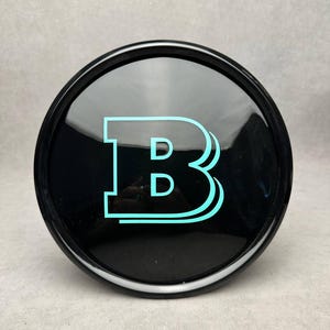 Puede incluir: Un emblema negro y redondo con un acabado brillante. Presenta la letra "B" estilizada, en color turquesa vibrante, con un sutil efecto de sombra. El diseño está centrado en el emblema.