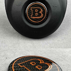Könnte beinhalten: Schwarze Airbag-Abdeckung für das Lenkrad mit einem orangefarbenen und schwarzen Emblem. Das Emblem zeigt den Buchstaben "B" mit zwei "D"s im Inneren. Das Emblem ist von einem Kohlefasermuster umgeben.
