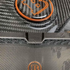 Könnte beinhalten: Nahaufnahme einer Kohlefaser-Oberfläche mit einem orange-schwarzen Emblem. Das Emblem zeigt den Buchstaben "B" in einem stilisierten Design. Die Kohlefaser hat ein gewebtes Muster, und das Emblem ist kreisförmig.