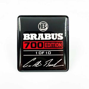 Puede incluir: Un emblema cuadrado negro brillante con el texto "BRABUS 700 EDITION" en blanco y rojo. El emblema también presenta el número "1 OF 10" y una firma en blanco. Un logotipo "B" blanco en la parte superior.