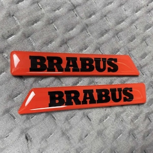 May include: Two orange rectangular emblems with the word "BRABUS" in bold black letters. The emblems have a glossy finish and are set against a textured grey background.