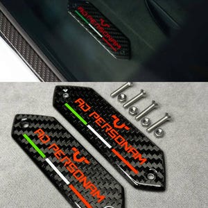 Op de afbeelding: Twee zeshoekige koolstofvezel plaatjes met de tekst AD PERSONAM in het rood, vergezeld van een groene, witte en rode streep. Elk plaatje heeft twee montagegaten en wordt geleverd met vier zilverkleurige metalen schroeven en vier moeren.