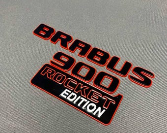 Può includere: Emblema BRABUS 900 ROCKET EDITION nero e rosso. Il testo è in grassetto, senza grazie, con un contorno rosso. L'emblema è su una superficie grigia.