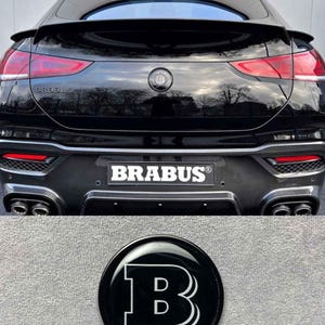 Peut inclure: Arrière d'une voiture noire avec feux arrière rouges et le mot "BRABUS" en blanc. Un emblème noir et blanc avec la lettre "B" est également visible. La voiture a un aileron et des doubles tuyaux d'échappement.