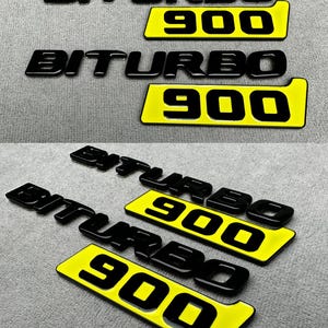 May include: Black "BITURBO" and "900" emblems with a glossy finish. The "900" is on a yellow background. The emblems are likely for automotive use.