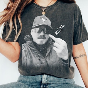 Op de afbeelding: Donkergrijs t-shirt met een zwart-wit afbeelding van een man met een pet en zonnebril, die de middelvinger opsteekt. Het shirt heeft een versleten uitstraling en een handtekening aan de rechterkant.