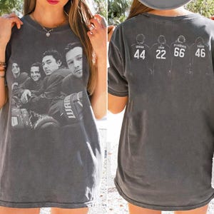 Puede incluir: Una camiseta gris carbón de manga corta con una impresión fotográfica en blanco y negro de cuatro jugadores de hockey en el frente. La parte posterior muestra cuatro siluetas blancas de jugadores con los nombres Graham 44, Logan 22, Di Laurentis 66 y Tucker 46 impresos debajo de cada figura.