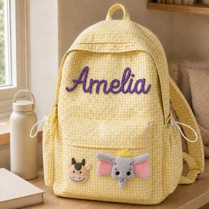 May include: A yellow and white gingham backpack featuring the name Amelia in purple cursive lettering. The front pocket is decorated with two plush appliques: a small brown cow face and a grey elephant with large pink ears and a yellow crown.