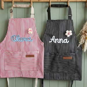 Puede incluir: Dos delantales a rayas colgando de un perchero de madera. Uno es rojo y blanco con el nombre "Olivia" en escritura azul y un pin de conejito. El otro es blanco y negro con "Anna" en escritura blanca y un pin de flor. Ambos tienen bolsillos.
