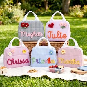 May include: Five rectangular lunch bags with white handles and striped fabric in black, light purple, pink, light blue, and light pink. Each bag features a unique name in raised, colorful yarn lettering and small crocheted floral or butterfly accents.