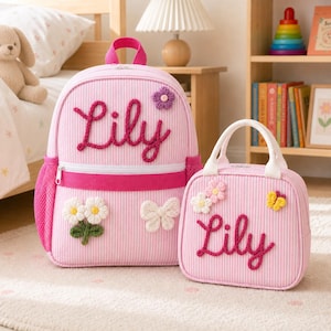 Op de afbeelding: Een roze en wit gestreepte rugzak en een bijpassende lunchtas, beide voorzien van de naam Lily in donkerroze garenletters. De rugzak heeft een paarse bloem, witte madeliefjes en een witte vlinder. De lunchtas heeft witte bloemen en een gele vlinder.