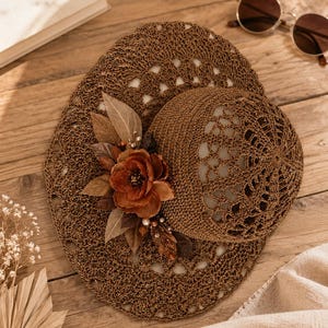 May include: A brown crocheted sun hat featuring an intricate openwork pattern throughout the crown and wide brim. The hat is decorated with a cluster of fabric leaves, a large rust-colored flower, and small pearl-like beads on the side.