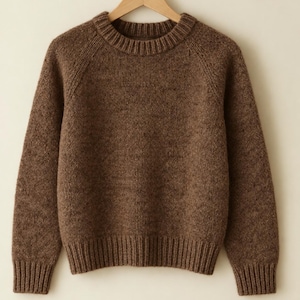 May include: A brown long-sleeve knit sweater with a crew neckline, ribbed cuffs, and a ribbed hem. The sweater features a speckled texture throughout the fabric and is displayed on a wooden hanger. The word HANDMADE is printed below the sweater.