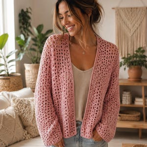 May include: A light pink open-front crochet cardigan featuring a textured, airy stitch pattern with long sleeves and a straight hemline, worn over a simple beige camisole and light blue denim jeans, showcasing a relaxed and comfortable fit.