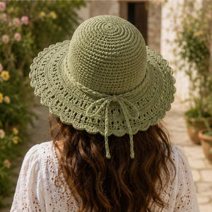 May include: A sage green crochet sun hat featuring a rounded crown and a wide, scalloped brim. The hat is accented with a matching crocheted cord tied into a bow at the back, showcasing intricate openwork patterns throughout the brim's design.