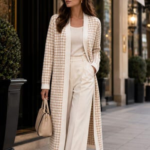May include: A long, open-front cardigan featuring a beige and white houndstooth pattern, worn over a cream-colored top and matching wide-leg trousers. The outfit is paired with a structured beige handbag and dark-tinted sunglasses.