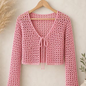 Peut inclure: Un cardigan à manches longues rose clair présentant un motif en crochet à mailles ouvertes avec une encolure en V et une fine fermeture à nouer sur le devant. Le vêtement est court et suspendu à un cintre en bois sur un fond neutre.