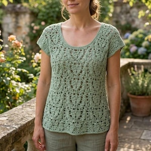 Può includere: Un top all'uncinetto verde chiaro con scollo tondo e maniche corte. Il top presenta un motivo traforato. La persona indossa pantaloncini verde chiaro. Lo sfondo comprende un muro di pietra e vegetazione.