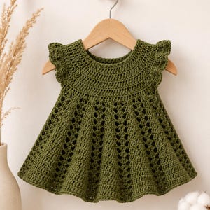 Könnte beinhalten: Ein olivgrünes Häkelkleid mit rundem Ausschnitt, gerüschten Flügelärmeln und einem ausgestellten Rock mit einem sich wiederholenden Rautenmuster. Das Kleidungsstück hängt auf einem hellen Holzkleiderbügel vor einem neutralen Hintergrund.