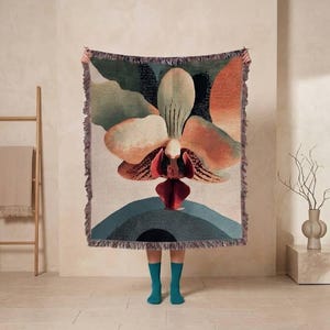 Op de afbeelding: Een geweven plaid met een grote, centrale orchidee in tinten perzik, oranje en donkerrood. Het ontwerp bevat abstract groen gebladerte en een donkerblauwe halfronde vorm aan de onderkant, afgewerkt met een gefranjerde rand.