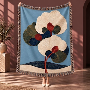 May include: A woven throw blanket featuring a light blue background with a large floral design. The pattern displays two prominent cream-colored flowers with deep red centers and dark green foliage. The blanket has a decorative fringed edge.