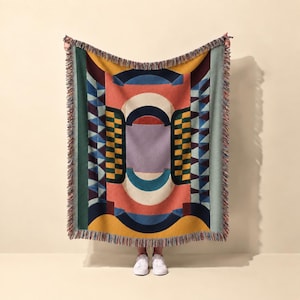 Puede incluir: Una manta tejida con un patrón geométrico que presenta un óvalo central color lavanda, rodeado por arcos concéntricos en crema, verde azulado y coral. El diseño incluye paneles laterales con motivos de tablero de ajedrez amarillo y verde oscuro.