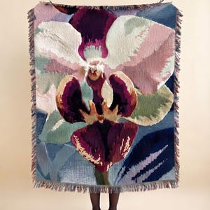 Puede incluir: Una manta tejida rectangular que presenta una gran flor de orquídea detallada en tonos púrpura intenso, rosa suave y blanco. La manta tiene un borde con flecos en todos los lados y muestra un patrón de tejido complejo y texturizado.