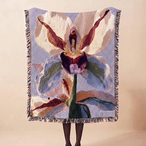 Puede incluir: Una manta tejida rectangular que presenta una orquídea grande y detallada en tonos crema, burdeos intenso y azul suave. El fondo tiene un patrón de cuadros finos en azul y blanco, y los bordes están terminados con un borde de flecos.