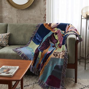 Puede incluir: Una manta tejida con un patrón abstracto y multicolor en tonos azul oscuro, morado, amarillo mostaza y verde claro. La manta rectangular tiene un borde con flecos de hilos multicolores y un acabado texturizado tipo tapiz.