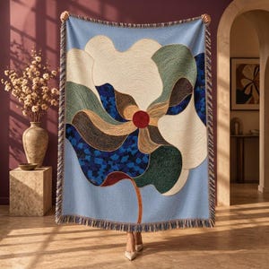 Puede incluir: Una manta tejida con un gran diseño floral abstracto en tonos azul claro, crema, verde oscuro, marrón y azul intenso, con un círculo rojo central. La manta rectangular tiene bordes con flecos en la parte superior e inferior.